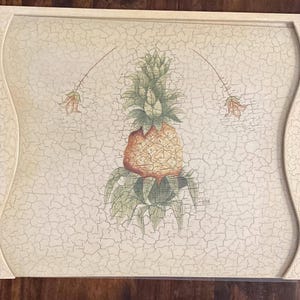 Puede incluir: Bandeja decorativa rectangular con acabado blanquecino envejecido. La bandeja presenta una ilustración central de piña con hojas verdes y una fruta naranja texturizada. Acentos florales y bordes curvos completan el diseño.