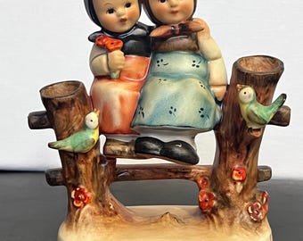 Könnte beinhalten: Vintage Keramikfigur mit zwei Kindern auf einer Holzbank. Die Kinder tragen traditionelle Kleidung, eines hält eine Blume. Die Bank ist mit kleinen Vögeln und Blumen verziert. Der Sockel ist cremefarben.