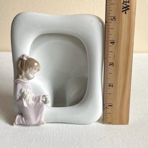Puede incluir: Un marco de fotos de cerámica blanca con una figura arrodillada con una túnica rosa claro que sostiene un pequeño ramo. Una regla de madera Westcott está colocada verticalmente junto al marco, mostrando una altura de unos 15 cm para el objeto.