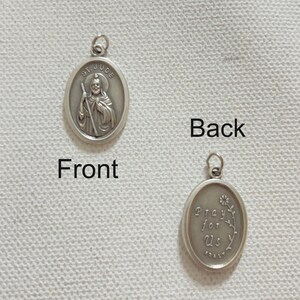 Op de afbeelding: Dubbelzijdige zilverkleurige St. Jude hanger. De voorkant toont een afbeelding van St. Jude met de tekst "ST. JUDE". De achterkant heeft de tekst "Pray for Us" en "ITALY" met een bloemenontwerp. De hanger is ovaal van vorm.