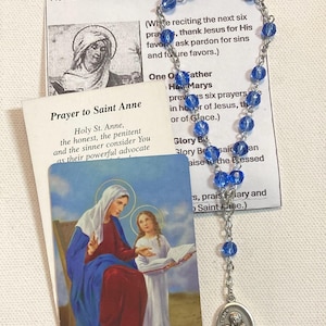 May include: A blue beaded rosary with a silver chain and a Saint Anne medal. Includes prayer cards with text about Saint Anne and the Chaplet Prayers. The cards feature illustrations of Saint Anne and the Virgin Mary.