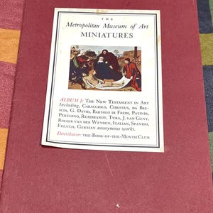 May include: A maroon book with a white label featuring the text "The Metropolitan Museum of Art MINIATURES." The label includes an image of a religious painting and text listing artists and the distributor, "THE-BOOK-OF-THE-MONTH CLUB."