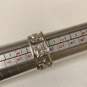 May include: A silver-toned ring featuring a repeating pattern of embossed angel figures. The ring is positioned on a metal ring sizing mandrel, with the band resting between the size 7 and 8 markings on the measurement scale.