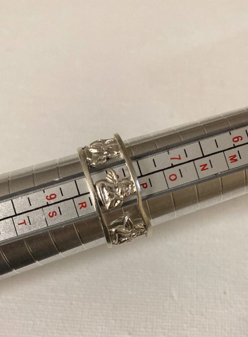 May include: A silver-toned ring featuring a repeating pattern of embossed angel figures. The ring is positioned on a metal ring sizing mandrel, with the band resting between the size 7 and 8 markings on the measurement scale.