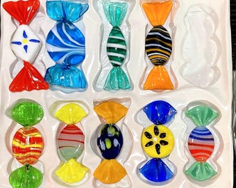 Könnte beinhalten: Eine Sammlung bunter Glasbonbon-Ornamente in einer weißen Schale. Die Bonbons weisen verschiedene Designs auf, darunter Streifen, Wirbel und florale Muster, mit leuchtenden Farben wie Rot, Blau, Grün, Gelb und Orange. Jedes Stück ist einzeln verpackt.