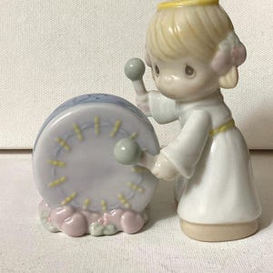May include: A porcelain figurine of an angel with blonde hair and a yellow halo, wearing a white robe with a yellow belt. The angel is holding two light green drumsticks and is positioned next to a round, light blue drum with yellow accents.