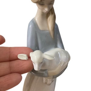 K&ouml;nnte beinhalten: Porzellanfigur einer Frau, die ein Lamm h&auml;lt. Die Frau tr&auml;gt ein hellblaues Kleid und hat blondes Haar. Das Lamm ist wei&szlig;. Ein kleines St&uuml;ck der Figur ist abgebrochen.