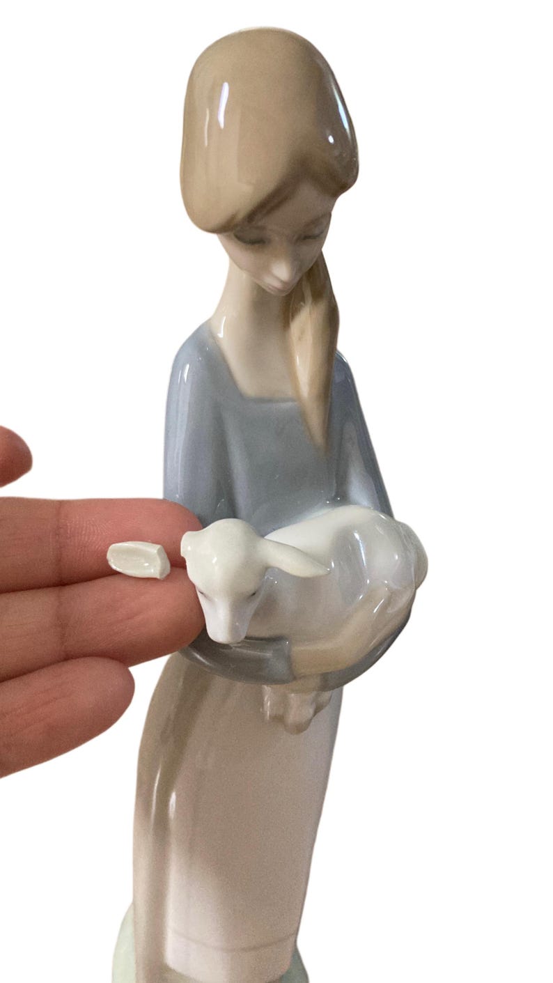 K&ouml;nnte beinhalten: Porzellanfigur einer Frau, die ein Lamm h&auml;lt. Die Frau tr&auml;gt ein hellblaues Kleid und hat blondes Haar. Das Lamm ist wei&szlig;. Ein kleines St&uuml;ck der Figur ist abgebrochen.