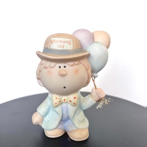 May include: A ceramic figurine of a young person wearing a hat that reads "BALLOONS 10¢". The figure is holding a bunch of pastel-coloured balloons. The figure is wearing a light blue jacket and a bow tie.