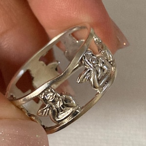 May include: A silver-toned metal ring featuring a cut-out design that depicts multiple small winged figures positioned around the band. The band has a smooth, polished finish and an openwork structure that creates negative space between each figure.