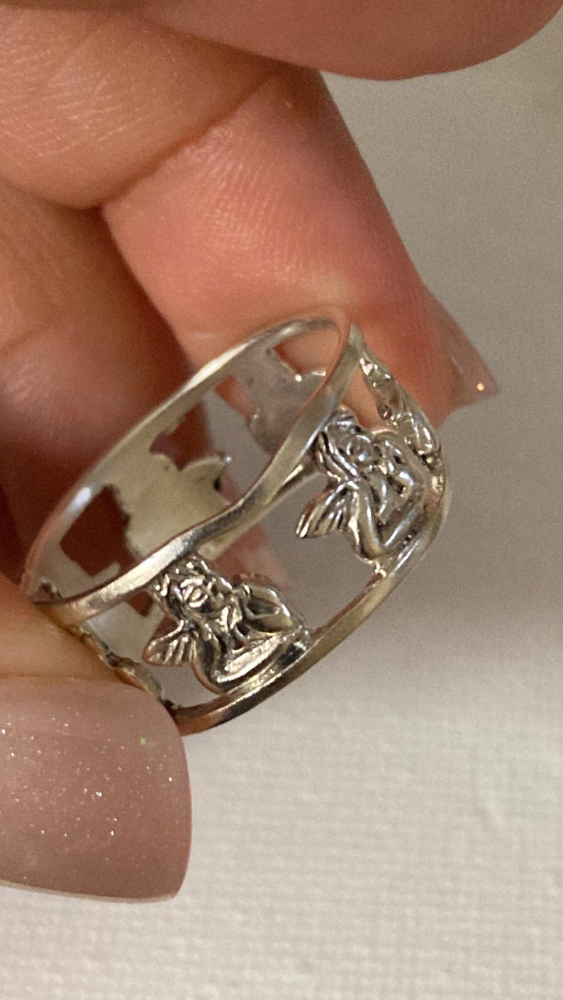 May include: A silver-toned metal ring featuring a cut-out design that depicts multiple small winged figures positioned around the band. The band has a smooth, polished finish and an openwork structure that creates negative space between each figure.