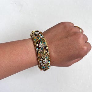 May include: A gold-toned hinged bangle bracelet featuring an intricate openwork vine and leaf design. The surface is embellished with numerous small, multi-colored iridescent rhinestones in shades of blue, green, orange, and clear, creating a textured, sparkling appearance across the entire outer band.