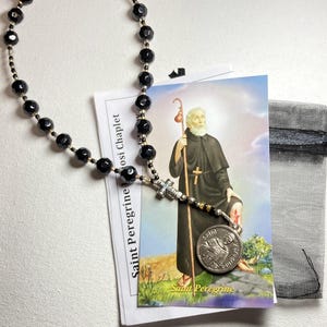 May include: A black beaded rosary with a silver cross and a Saint Peregrine card. The card features an illustration of Saint Peregrine and a silver coin with an image of Saint Peregrine.
