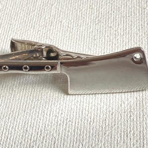 May include: A silver-colored tie clip in the shape of a cleaver. The clip has a shiny, reflective surface and features three small, round rivets on the handle. The cleaver blade has a single, round rivet on the end.