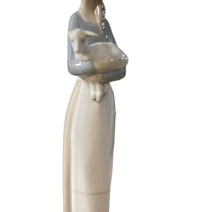 K&ouml;nnte beinhalten: Porzellanfigur einer Frau, die ein Lamm h&auml;lt. Die Frau tr&auml;gt ein langes, helles Kleid mit einem grauen Oberteil und ihr Haar ist zur&uuml;ckgebunden. Das Lamm ist wei&szlig; und wird in ihren Armen gehalten. Die Figur steht auf einem gr&uuml;nen Sockel.