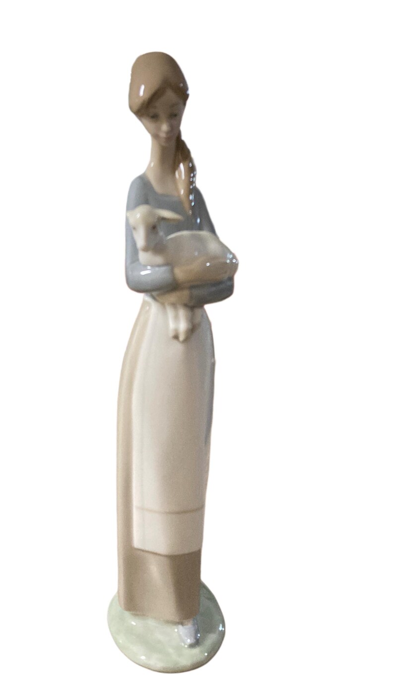 K&ouml;nnte beinhalten: Porzellanfigur einer Frau, die ein Lamm h&auml;lt. Die Frau tr&auml;gt ein langes, helles Kleid mit einem grauen Oberteil und ihr Haar ist zur&uuml;ckgebunden. Das Lamm ist wei&szlig; und wird in ihren Armen gehalten. Die Figur steht auf einem gr&uuml;nen Sockel.