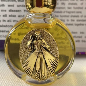 Puede incluir: Una pequeña botella redonda de vidrio con tapa dorada. La botella contiene un líquido amarillo y presenta un medallón dorado con una figura religiosa. La botella es probablemente para perfume o aceites esenciales.