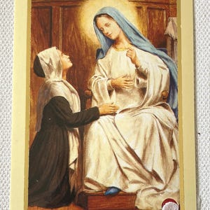 Op de afbeelding: Vintage religieuze kunstafdruk met een vrouw in een witte jurk en blauwe hoofdbedekking die op een stoel zit, met een vrouw in een zwarte jurk die voor haar knielt. De afbeelding heeft een gouden rand en een warm kleurenpalet.