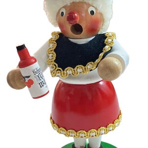 May include: Wooden figurine of a woman in traditional attire, holding a small bottle. The figure has a red hat, black vest with gold trim, and a red skirt. The bottle has text and cherry illustrations.