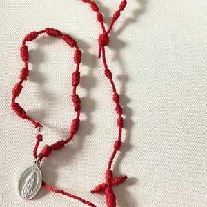May include: Red knotted rosary with a silver pendant featuring a religious figure. The rosary has a cross-shaped pendant and is made of red cord. The beads are knotted and evenly spaced.