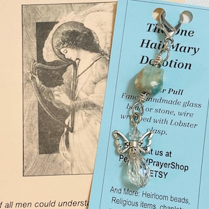 May include: A handmade angel charm with a clear crystal body and silver wings, attached to a blue-green stone bead. The charm is connected to a silver clasp. A blue tag with text about the charm and Etsy shop is in the background.