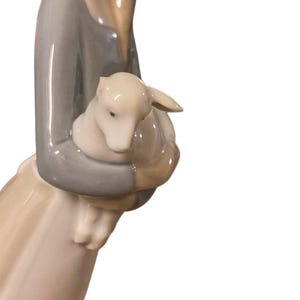 K&ouml;nnte beinhalten: Porzellanfigur einer Frau, die ein Lamm h&auml;lt. Die Frau tr&auml;gt ein graues Oberteil und einen langen Rock. Das Lamm ist wei&szlig;. Die Figur hat ein ruhiges und zartes Aussehen.