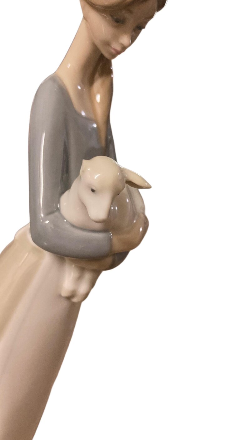 K&ouml;nnte beinhalten: Porzellanfigur einer Frau, die ein Lamm h&auml;lt. Die Frau tr&auml;gt ein graues Oberteil und einen langen Rock. Das Lamm ist wei&szlig;. Die Figur hat ein ruhiges und zartes Aussehen.