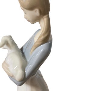 K&ouml;nnte beinhalten: Porzellanfigur einer jungen Person, die ein Lamm h&auml;lt. Die Figur tr&auml;gt ein hellblaues Oberteil und einen wei&szlig;en Rock. Das Lamm ist wei&szlig;. Die Figur hat einen ruhigen Ausdruck und ist ein dekorativer Gegenstand.