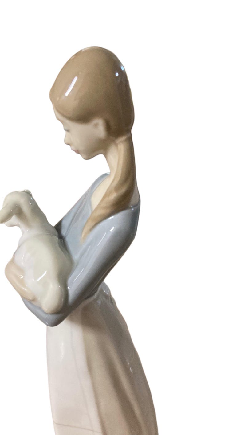 K&ouml;nnte beinhalten: Porzellanfigur einer jungen Person, die ein Lamm h&auml;lt. Die Figur tr&auml;gt ein hellblaues Oberteil und einen wei&szlig;en Rock. Das Lamm ist wei&szlig;. Die Figur hat einen ruhigen Ausdruck und ist ein dekorativer Gegenstand.