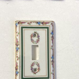 Könnte beinhalten: Eine rechteckige Lichtschalterabdeckung aus Keramik mit weißem Untergrund und einer zentralen Öffnung für den Kippschalter. Die Platte ist mit einem grünen rechteckigen Rahmen, Goldverzierungen und einer bunten Blumengirlande an den Rändern und um die Schraublöcher versehen.