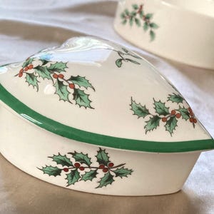 Op de afbeelding: Een hartvormige, crèmekleurige keramische sieradendoos met een groene rand en deksel. De doos is versierd met groene hulstbladeren en rode bessen. Een andere soortgelijke doos is zichtbaar op de achtergrond.