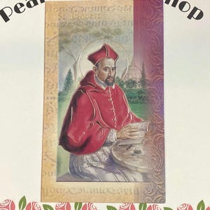 May include: A religious card featuring an illustration of St. Robert in a red robe and hat, holding a book. The background includes a church and trees. The text "Pearls of PrayerShop" is at the top.
