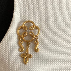 May include: A gold-colored brooch with an ornate design. The brooch features a central floral-like shape with dangling, curved elements and a cross-shaped pendant. It is pinned to a white textured fabric.