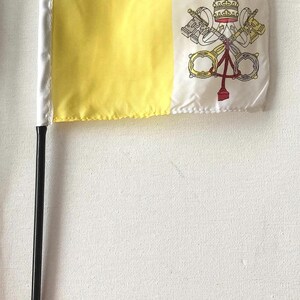 Puede incluir: Una pequeña bandera de la Ciudad del Vaticano con un diseño amarillo y blanco. La bandera presenta el escudo de armas del Vaticano en el lado blanco y está unida a un mástil negro con una punta de lanza dorada.