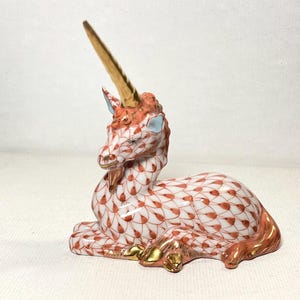 May include: A ceramic unicorn figurine in a resting pose. The white body is decorated with a red leaf pattern. The mane, tail, and hooves are accented with gold. The unicorn has a gold horn and blue ears.
