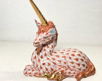 Könnte beinhalten: Keramik-Einhornfigur in Ruheposition. Der weiße Körper ist mit einem roten Blättermuster verziert. Mähne, Schwanz und Hufe sind mit Gold akzentuiert. Das Einhorn hat ein goldenes Horn und blaue Ohren.