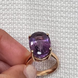 May include: A gold ring featuring a large, oval-cut purple amethyst gemstone set in a four-prong mounting. The band is smooth and polished, showcasing a warm metallic tone that complements the deep violet hue of the faceted central stone.