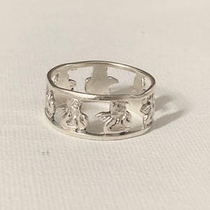 May include: A silver-toned band ring featuring a repeating cutout design of small winged figures. The ring has a polished metallic finish and a continuous openwork pattern that encircles the entire circumference of the circular band.