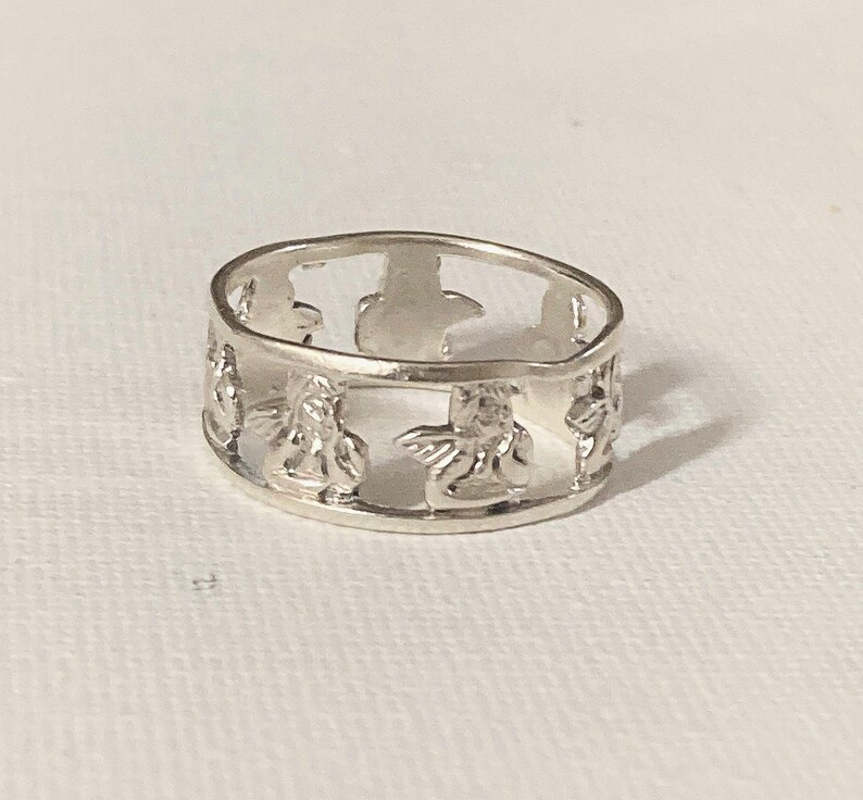 May include: A silver-toned band ring featuring a repeating cutout design of small winged figures. The ring has a polished metallic finish and a continuous openwork pattern that encircles the entire circumference of the circular band.