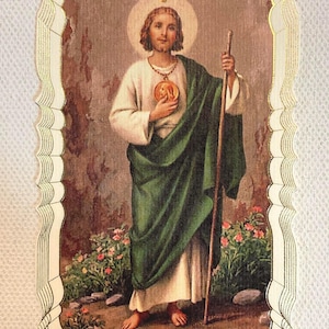 Op de afbeelding: Een religieuze afbeelding van een figuur in witte en groene gewaden, met een staf en een medaillon. De afbeelding heeft een goudkleurige, geschulpte rand. De achtergrond toont bloemen en een stenen muur.