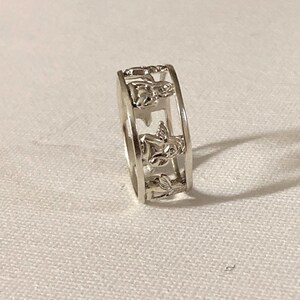 May include: A silver-toned metal ring featuring a cut-out design that displays a series of repeating cherub or angel figures around the entire circumference of the band. The ring has a polished finish and a consistent width throughout its structure.