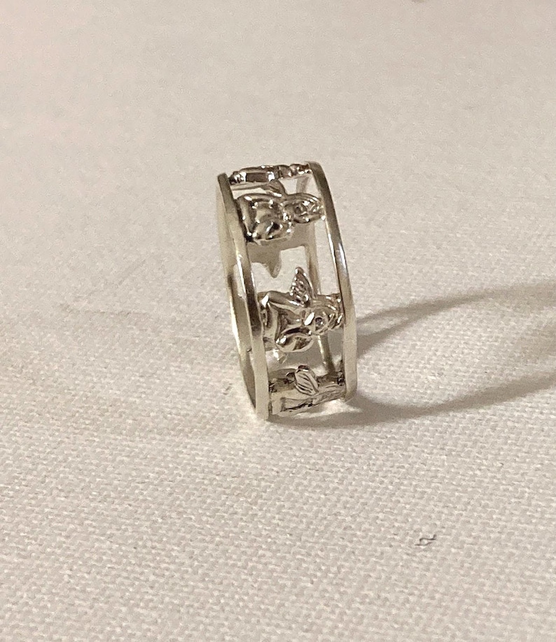May include: A silver-toned metal ring featuring a cut-out design that displays a series of repeating cherub or angel figures around the entire circumference of the band. The ring has a polished finish and a consistent width throughout its structure.