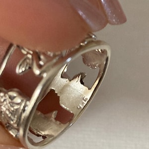 May include: A silver-toned ring featuring a cut-out design of a winged figure and floral motifs. The interior of the band is clearly stamped with the hallmark 925, indicating the metal composition. The ring has a polished finish and openwork.