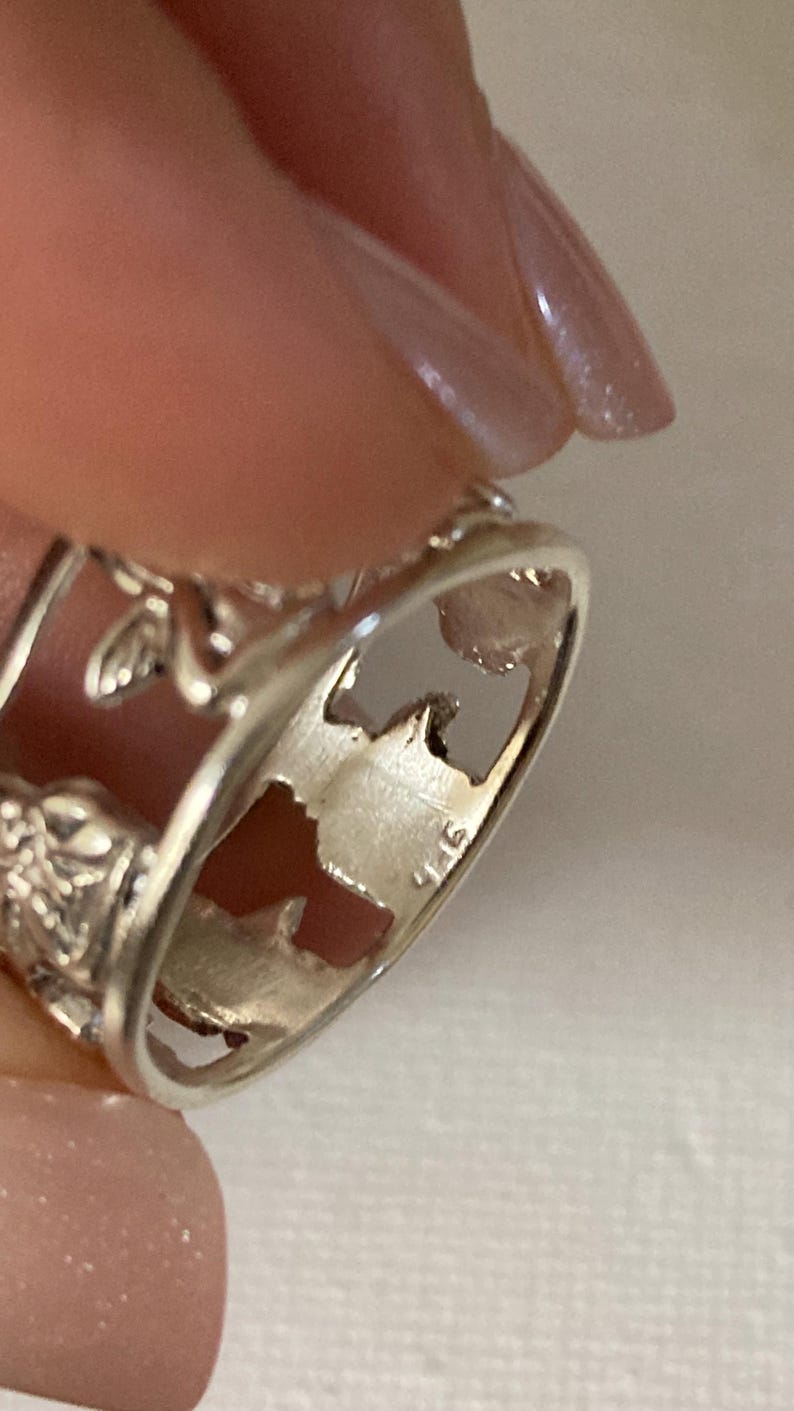 May include: A silver-toned ring featuring a cut-out design of a winged figure and floral motifs. The interior of the band is clearly stamped with the hallmark 925, indicating the metal composition. The ring has a polished finish and openwork.