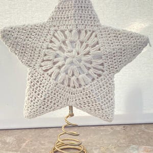 May include: A cream-colored, crocheted star tree topper with a floral design in the center. The star is mounted on a gold-colored metal spiral base. The star is a festive decoration for the holiday season.
