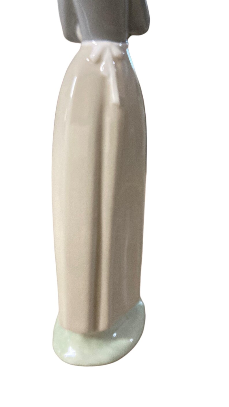 K&ouml;nnte beinhalten: Porzellanfigur einer Frau, von hinten gesehen. Die Figur tr&auml;gt ein langes, hellbeige Kleid mit einer wei&szlig;en Sch&uuml;rze, die an der Taille gebunden ist. Kopf und Schultern sind grau, der Sockel hellgr&uuml;n.