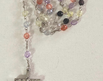 Könnte beinhalten: Ein Rosenkranz mit bunten Perlen in Orange, Lila, Gelb und Schwarz, auf einer silbernen Kette aufgereiht. Der Rosenkranz hat ein silbernes Kruzifix am unteren Ende und einen dekorativen Verschluss oben.