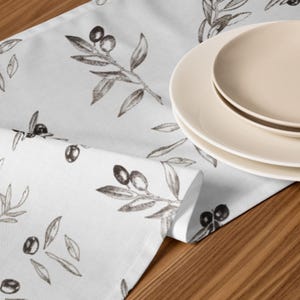 May include: A white table runner featuring a repeating dark gray sketch-style pattern of olive branches with leaves and olives. The fabric is folded over, and a stack of three plain, light cream-colored round ceramic plates rests on the runner.