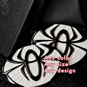 May include: Two oval-shaped enamel pins featuring a black spider emblem centered over a white web pattern. The text Any color, Any size, Any design is printed in red on the right side. The pins have a glossy finish and black outlines.