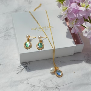 Könnte beinhalten: Ein goldenes Halsketten- und Ohrring-Set mit Perlen- und schillernden Abalone-Muschel-Anhängern. Die Ohrringe und der Anhänger haben ein Sonnenstrahl-Design. Das Schmuckstück wird auf einer weißen Schachtel mit dem Text "AQUA & PEARL LONDON" präsentiert.